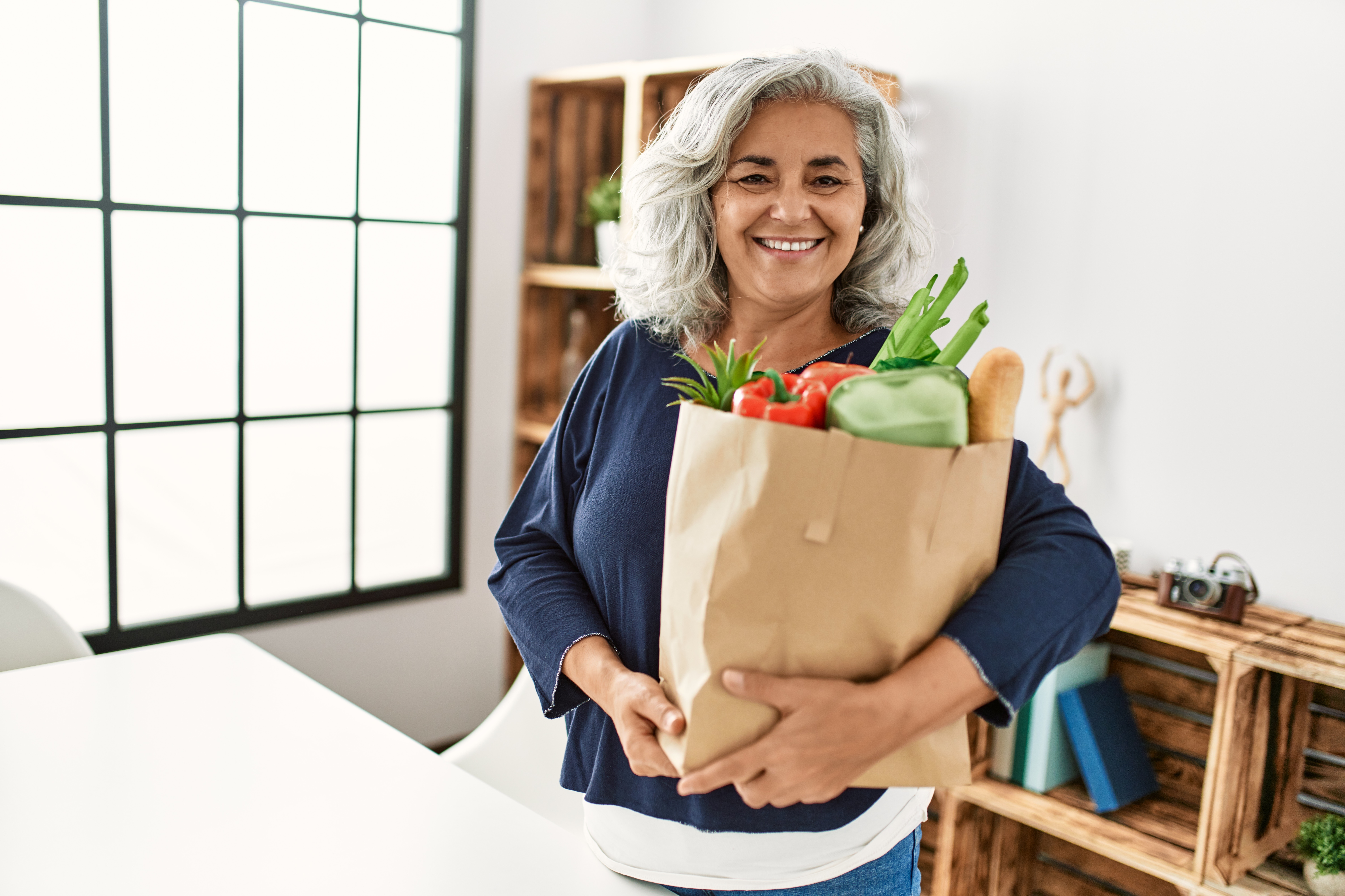 Middle age grey-haired woman holding paper bag with groceries standing at home.
2158795945